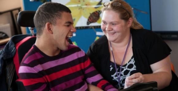 Young man in wheelchair smiles next to direct support professional
