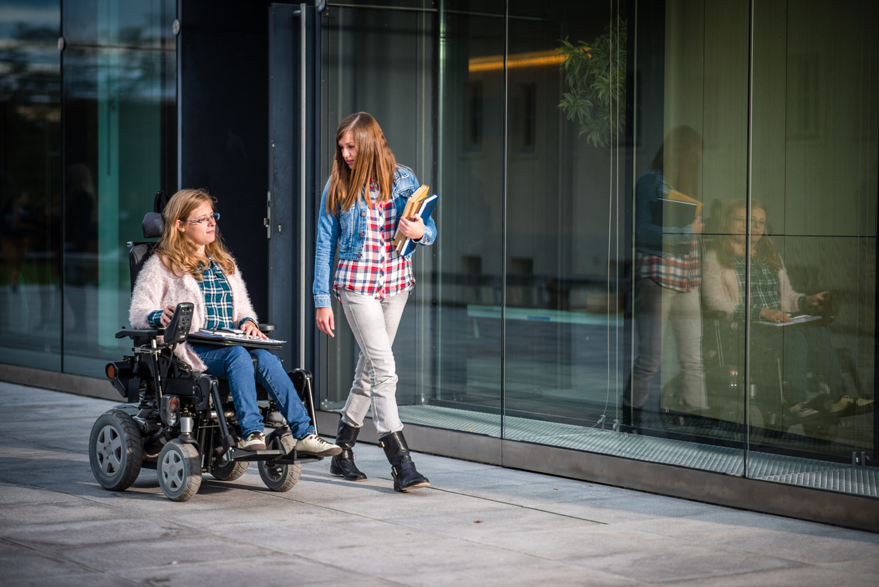 Two female college students, one using a wheelchair, walk side by side down a college campus.