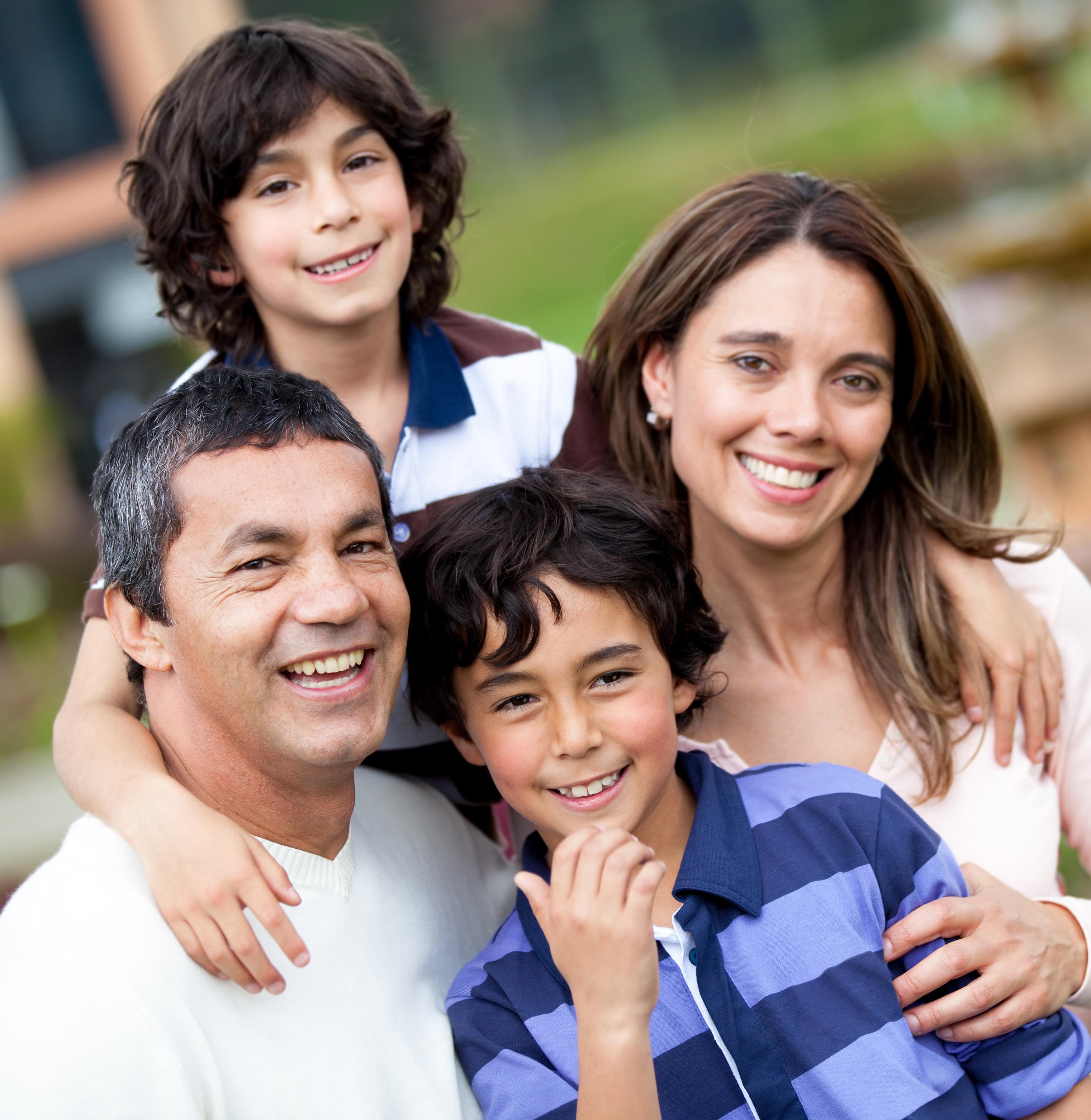 A family photo of four taken outside: a father and his wife are smiling and wearing white, one son in front is smiling and has a purple striped shirt on, one son in back is smiling, with his arm on his dad, has a blue and white striped shirt