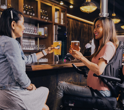 Two women share a drink at a bar, both are sitting in wheelchairs.